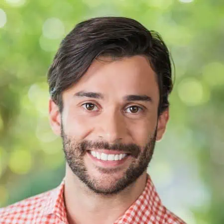 Friendly man with dark hair and beard smiling in natural outdoor setting.