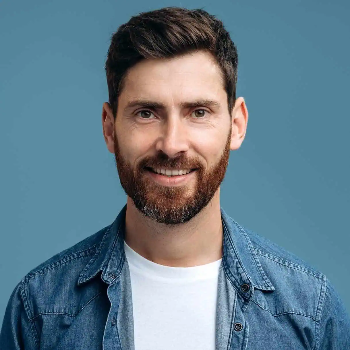 Man smiling with a beard, wearing a denim jacket against a blue background.