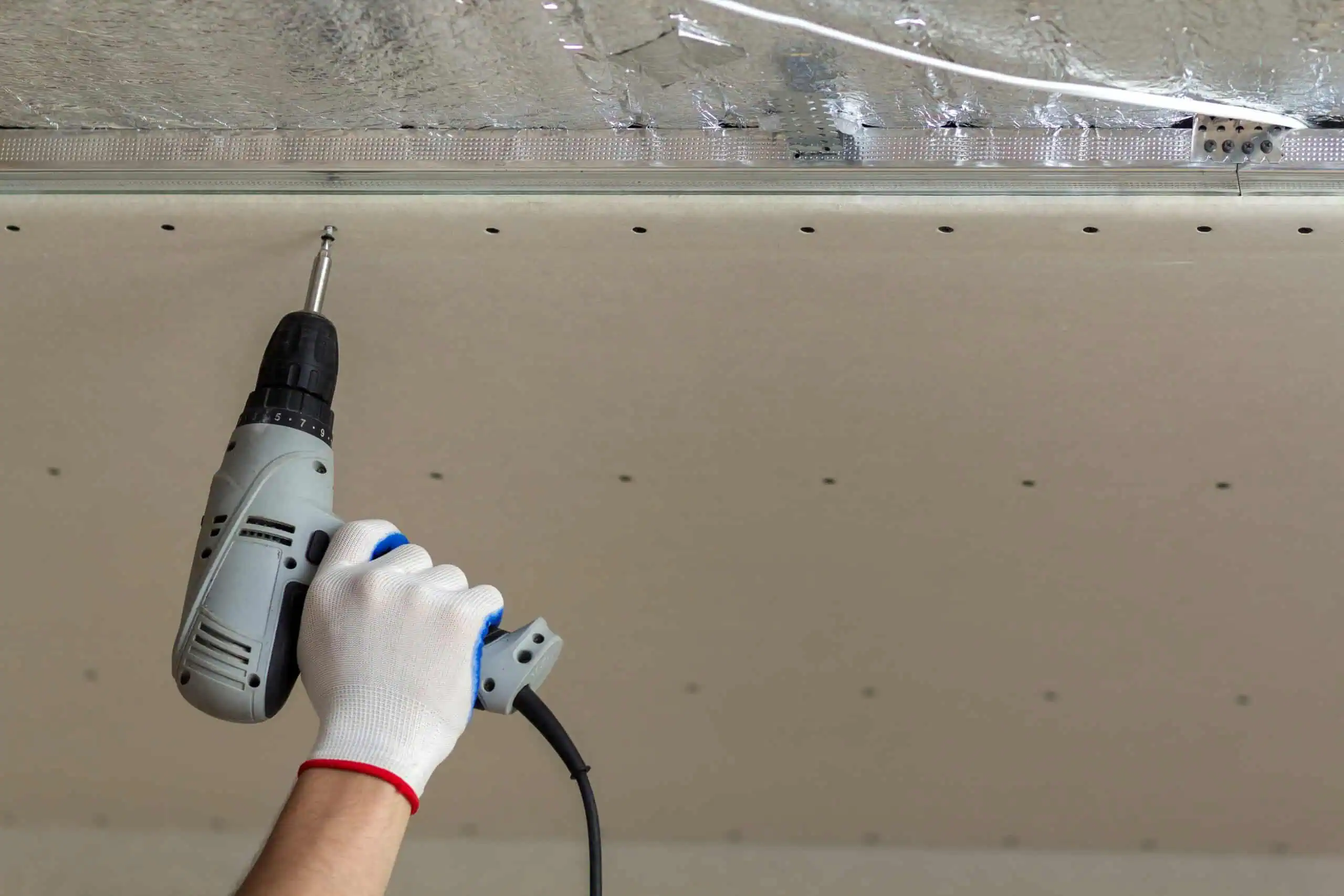 Person installing drywall with a power drill on ceiling.