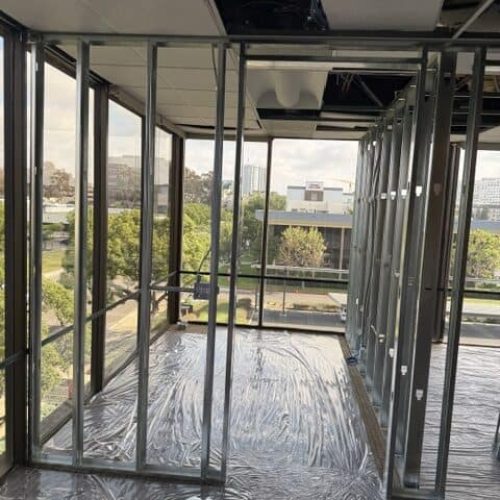 Drywall installation underway in a commercial building with large windows and metal framing.