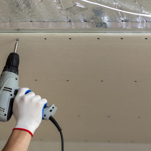 Close-up of constructor worker hand in protective glove with electrical cordless screwdriver connecting suspended drywall ceiling to metal frame. Renovation, construction and DIY concept.