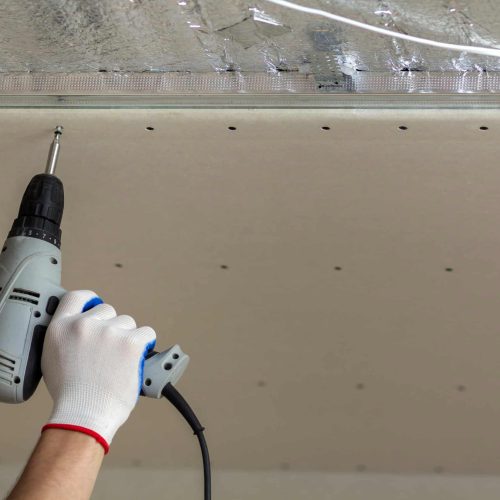 Person installing drywall with a power drill on ceiling.